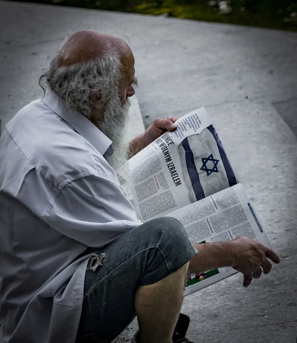 White-bearded man reading a newspaper with an Israeli flag on the page